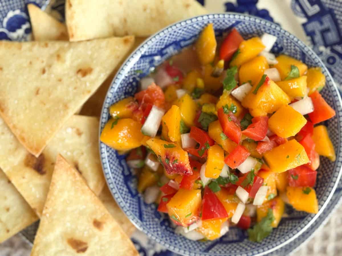 A blue and white bowl holds mango salsa with diced mango, red bell pepper, onion, and cilantro. Next to the bowl are several triangular baked tortilla chips on a patterned plate.