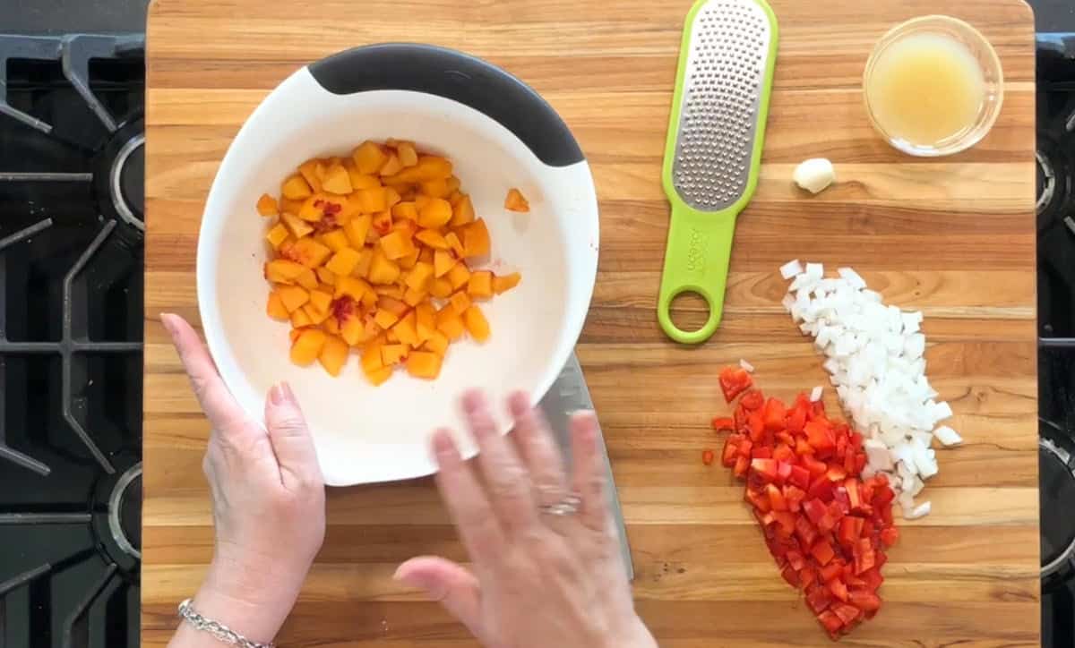 A person holds a bowl of chopped peaches over a wooden cutting board with diced onions, red bell pepper, a grater, a garlic clove, and a small bowl of liquid. A stove is visible to the left.