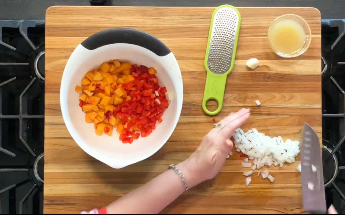 A person chops white onion on a wooden cutting board. Diced red and yellow bell peppers are in a mixing bowl nearby, with a hand grater, a garlic clove, and a small bowl of liquid beside them.