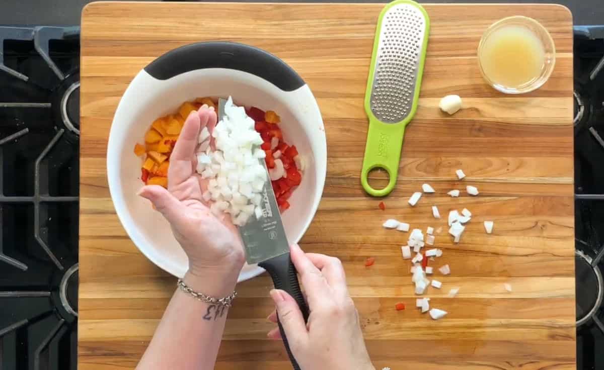 A person holds diced onions on a knife over a white bowl containing chopped yellow and red peppers on a wooden cutting board. A grater, garlic clove, and small bowl of liquid are nearby.