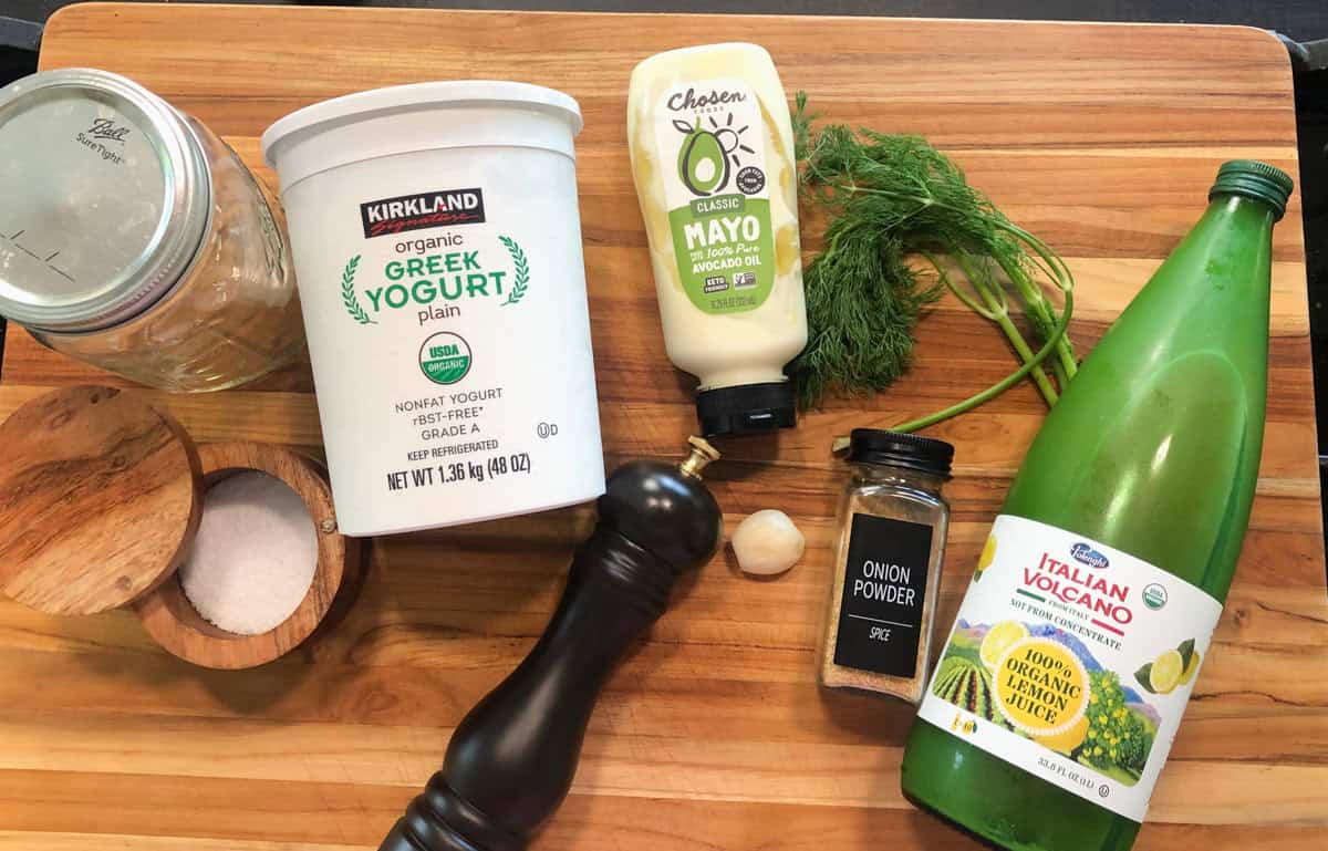 A wooden cutting board holds a jar of salt, a container of Greek yogurt, a black pepper grinder, mayonnaise, fresh dill, a garlic clove, onion powder, and a bottle of lemon juice.