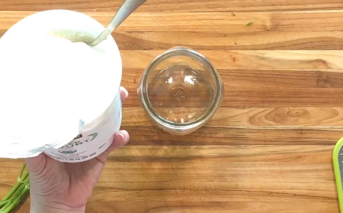 A hand holds a container of plain yogurt with a spoon inside, positioned next to an empty glass jar on a wooden countertop.