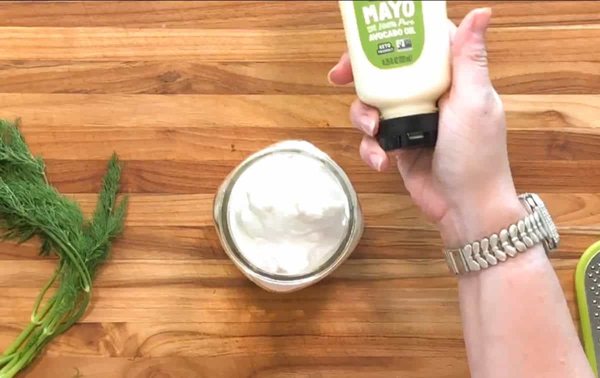 A hand holding a squeeze bottle of avocado oil mayonnaise over an open glass jar filled with a creamy white substance on a wooden surface. Fresh dill and a grater are also visible.