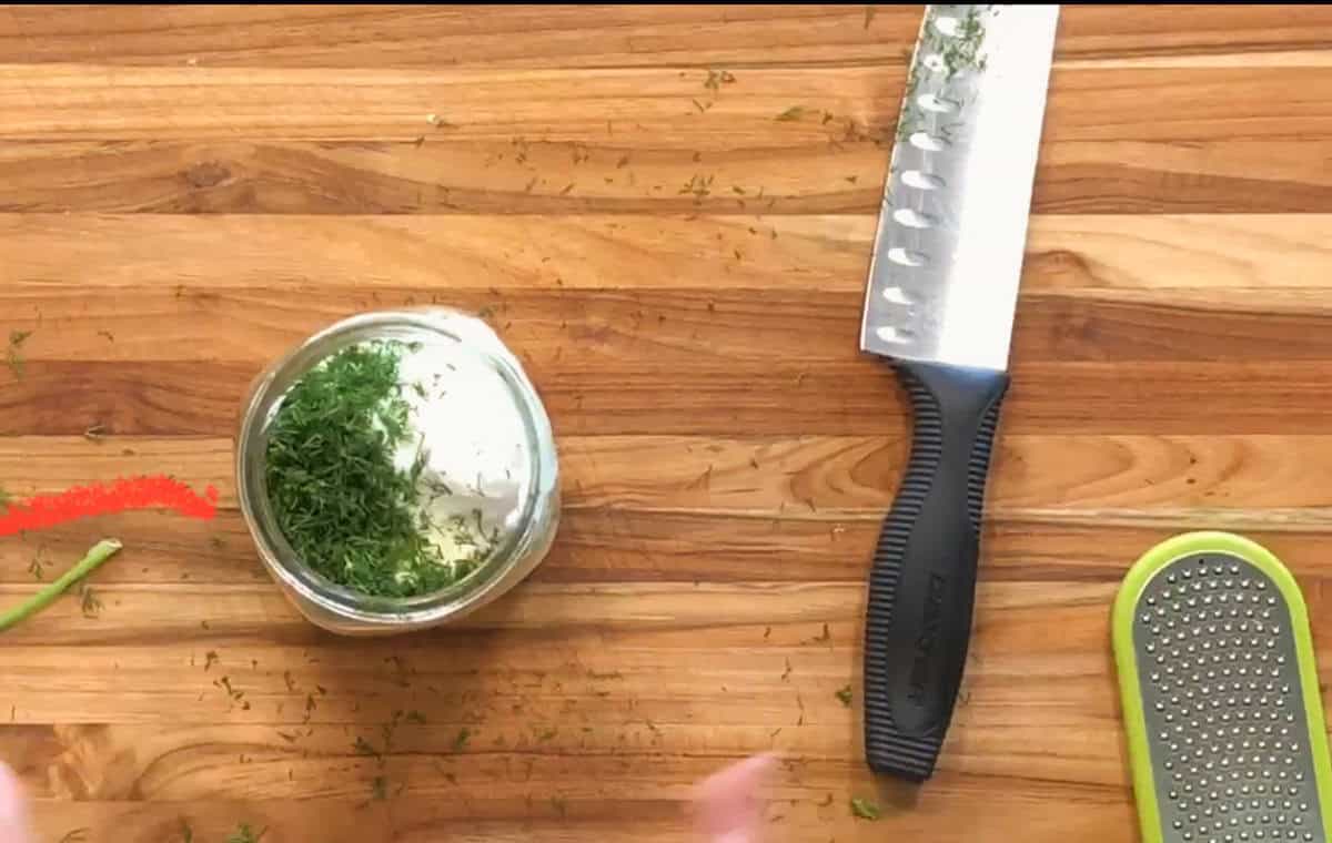 A wooden cutting board with a knife, a metal grater, and a small jar filled with herbs and a creamy substance. Some chopped herbs and a green stem are scattered on the board.