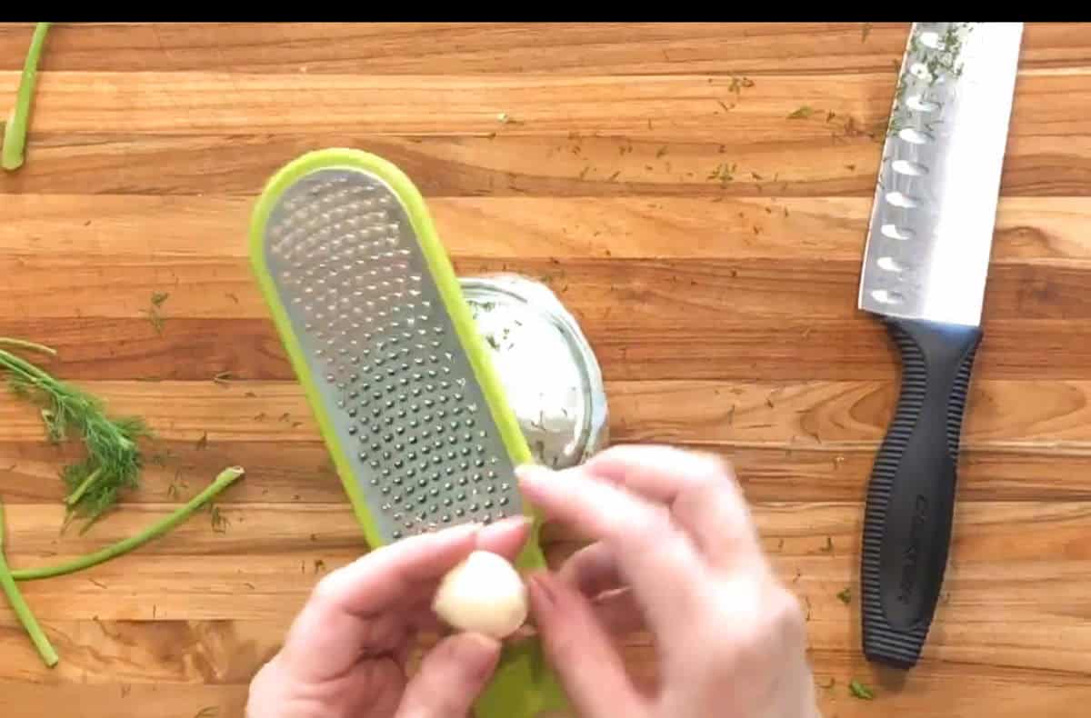 Two hands hold a clove of garlic over a grater positioned above a glass jar on a wooden countertop. A chef’s knife and some chopped herbs are also visible on the surface.