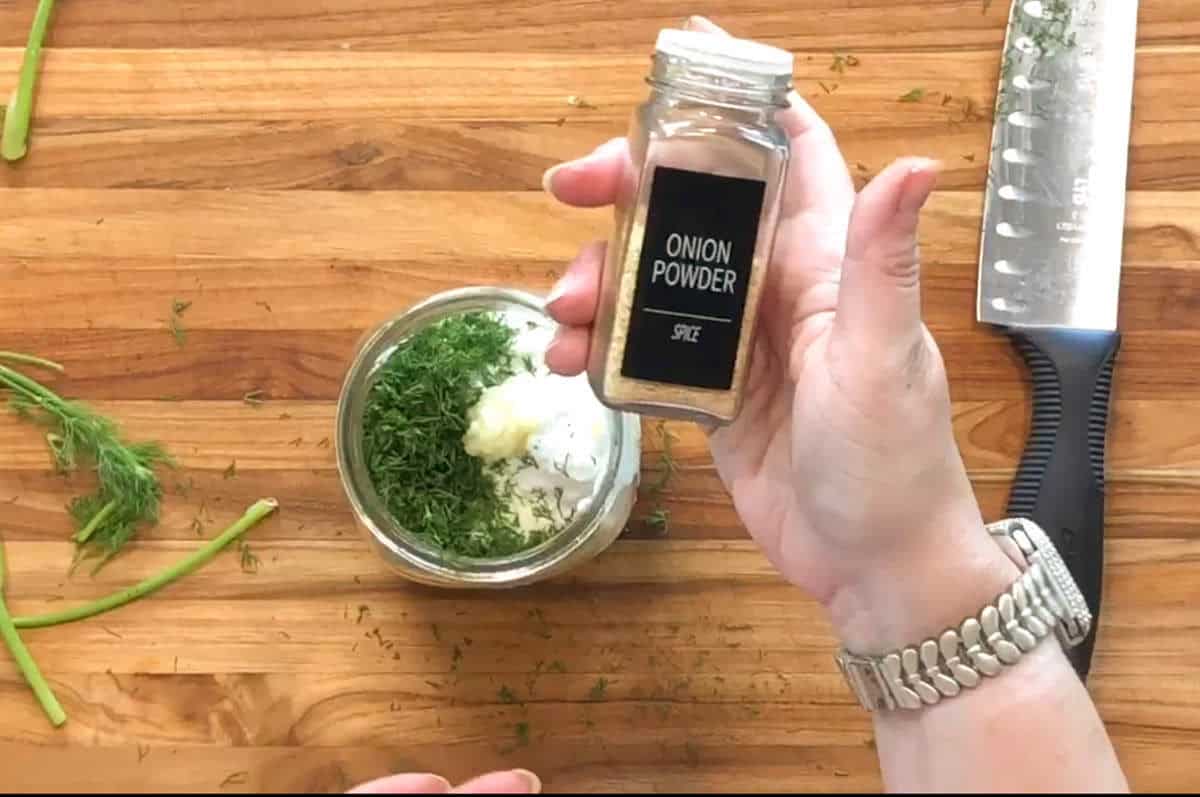 A hand holds a jar labeled "Onion Powder" above a bowl with chopped herbs, mayonnaise, and other ingredients. A large knife and herb stems are on a wooden cutting board.