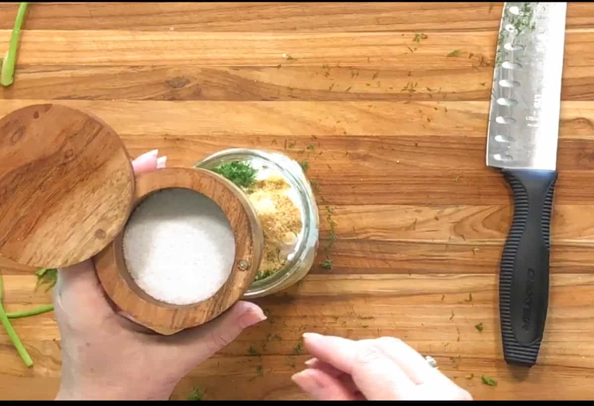 A hand holds an open wooden salt cellar above a glass jar containing mixed seasoning ingredients on a wooden cutting board. A chef’s knife rests to the right on the board.