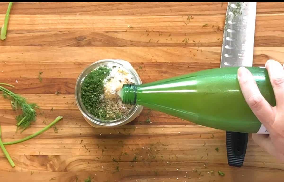 A hand pours liquid from a green bottle into a glass jar filled with chopped herbs and other ingredients on a wooden cutting board, next to a knife and herb stems.
