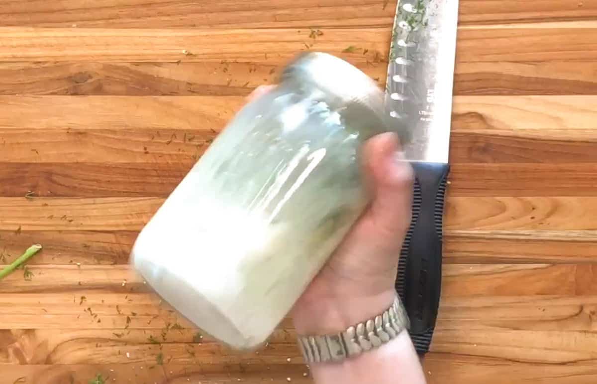 A hand wearing a watch is holding and shaking a closed glass jar with a creamy white mixture inside. The background is a wooden surface with a large knife and chopped herb pieces scattered around.