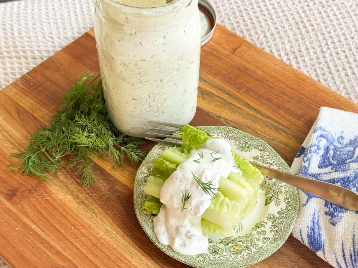A jar of creamy dill dressing sits on a wooden board next to fresh dill. A small plate holds celery and lettuce topped with the dressing. A fork and a blue-and-white cloth are nearby.