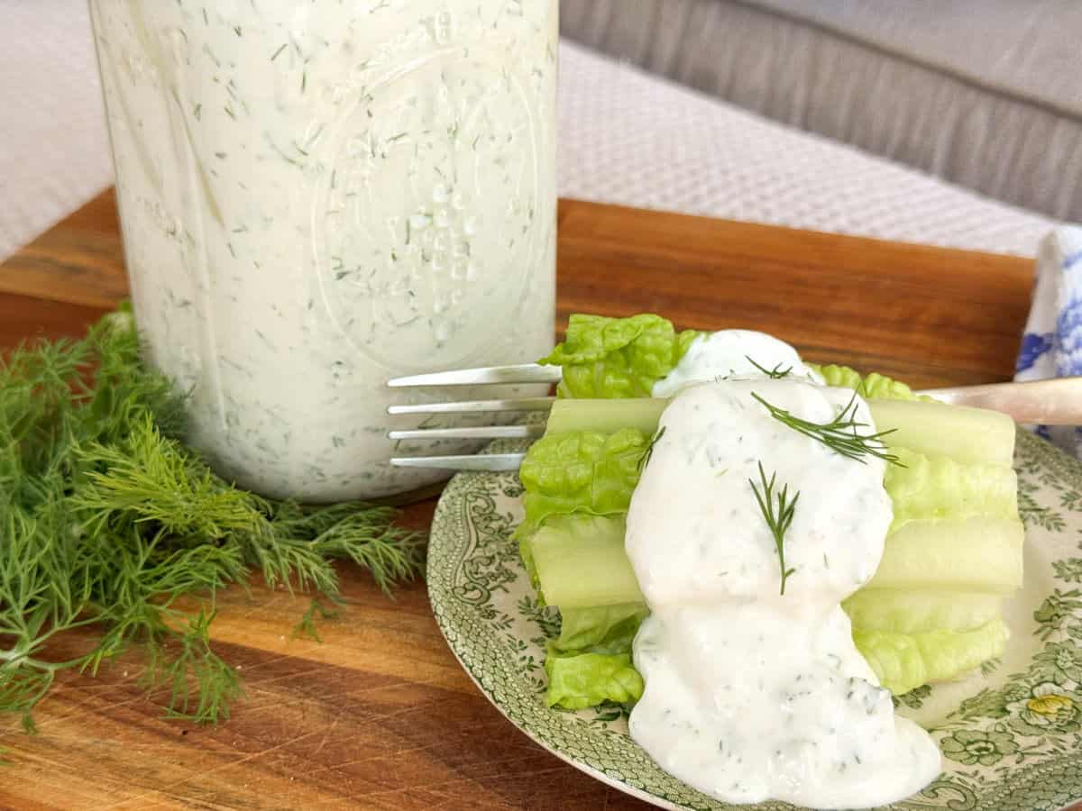 A plate with romaine lettuce and celery sticks topped with creamy dressing and dill, placed next to a fork, a bunch of fresh dill, and a jar filled with similar dressing, on a wooden surface.