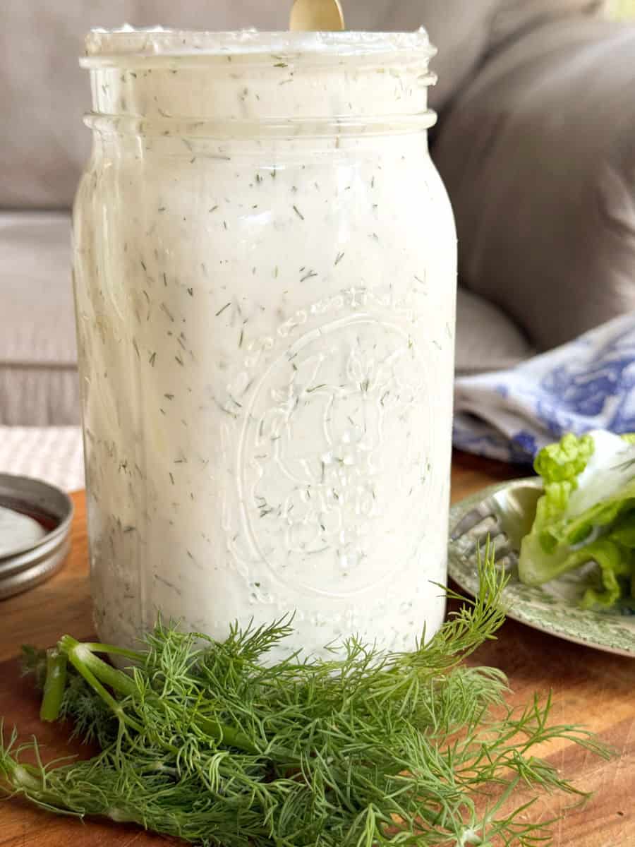 A glass jar filled with creamy white ranch dressing with visible dill, sitting on a wooden surface next to fresh dill and lettuce. A spoon is in the jar and a lid is nearby. A couch and towel are in the background.