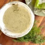A bowl of creamy herb dressing with a spoon inside, sitting on a wooden surface next to fresh dill and a plate with lettuce and a fork.