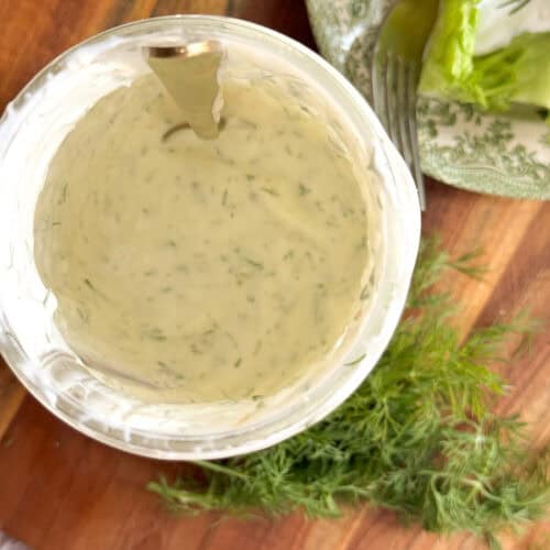 A bowl of creamy herb dressing with a spoon inside, sitting on a wooden surface next to fresh dill and a plate with lettuce and a fork.
