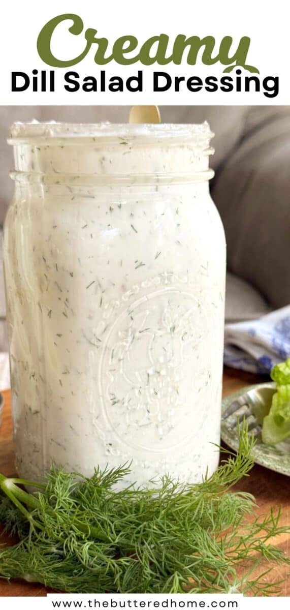 A large glass jar filled with creamy dill salad dressing sits on a wooden surface. Fresh dill is placed in front of the jar, and there are some lettuce leaves and a blue patterned cloth in the background.