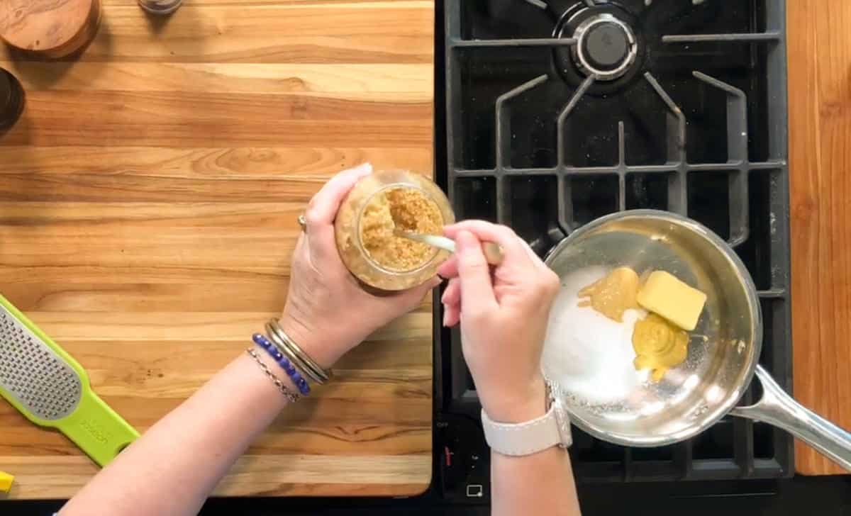 A person stirs brown sugar in a jar above a stove. On the stovetop, a saucepan contains brown sugar, butter, white sugar, and mustard, ready to be mixed. A grater lies on the wooden counter nearby.