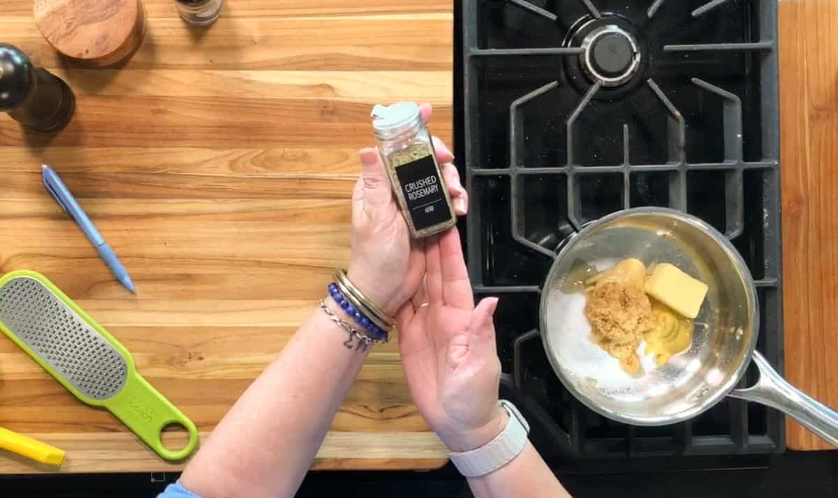 A person holds a jar labeled "ground tarragon" over a saucepan on a stove containing butter and brown sugar. A grater, pen, pepper mill, and spice jar sit on a wooden countertop nearby.