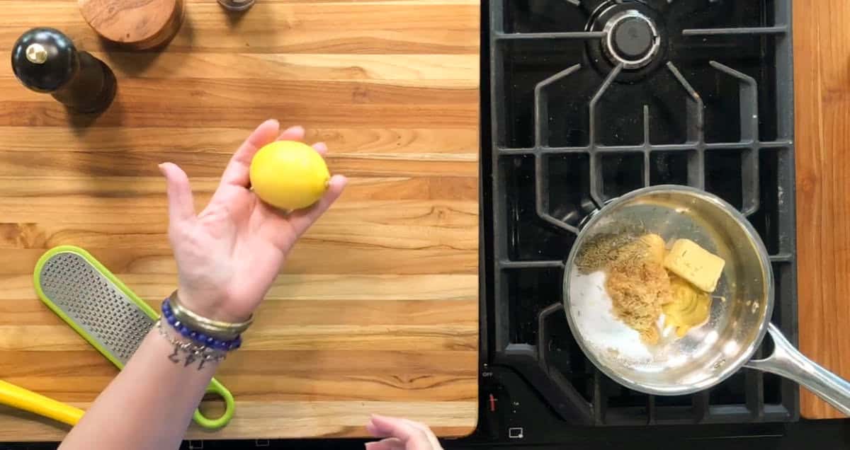 A person holds a lemon above a wooden countertop next to a grater. Beside them is a stovetop with a pan containing butter and seasonings. A pepper mill and a small bowl are also on the counter.