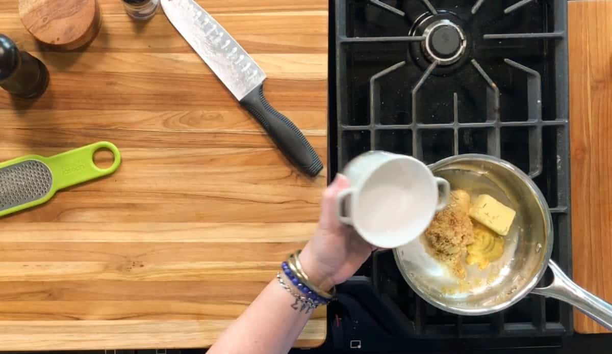 A person pours a white liquid from a small bowl into a saucepan containing butter and brown sugar on a stovetop, with a wooden cutting board, grater, knife, and pepper grinder nearby.