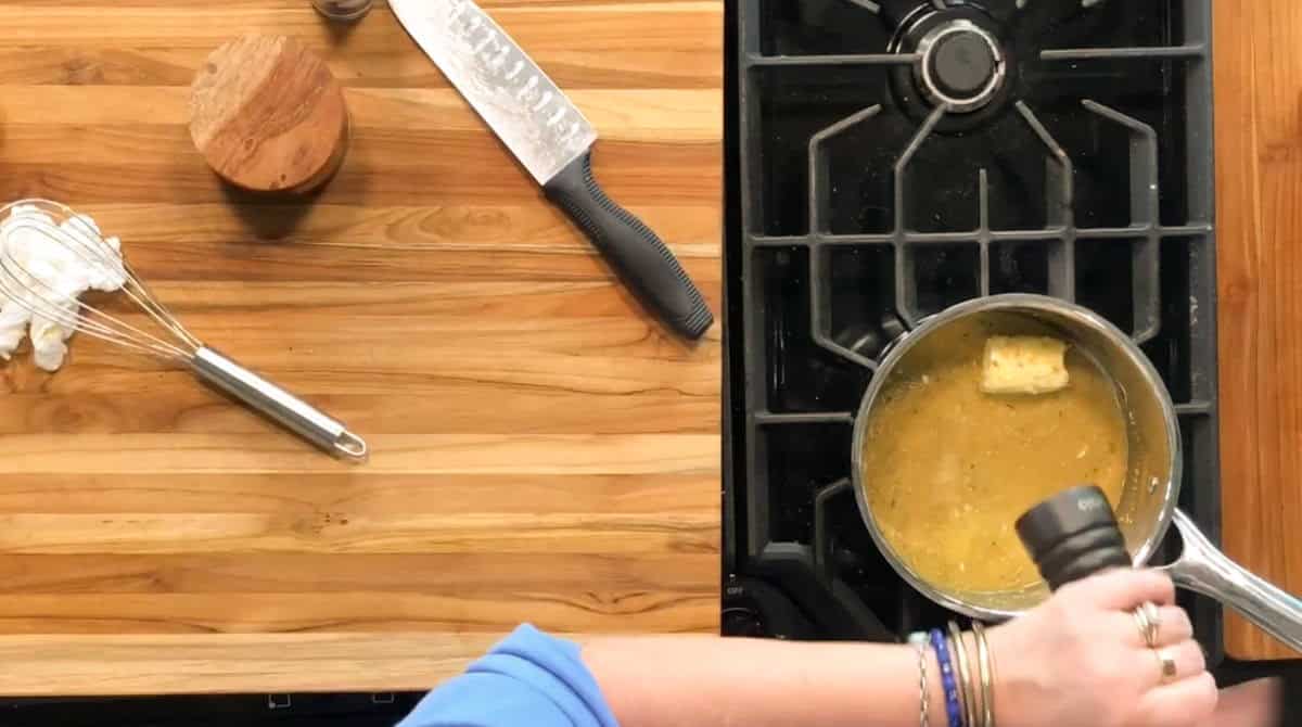 A person grinds black pepper into a saucepan with brown sauce and a pat of butter on a stove. A whisk, chef’s knife, small wooden container, and towel are on a wooden countertop beside the stove.