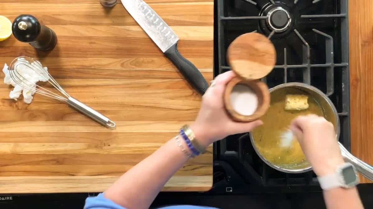 Person stirs food in a pot on a stove with one hand while holding an open salt container over the pot with the other; a large knife, whisk, and pepper grinder are on a wooden countertop nearby.