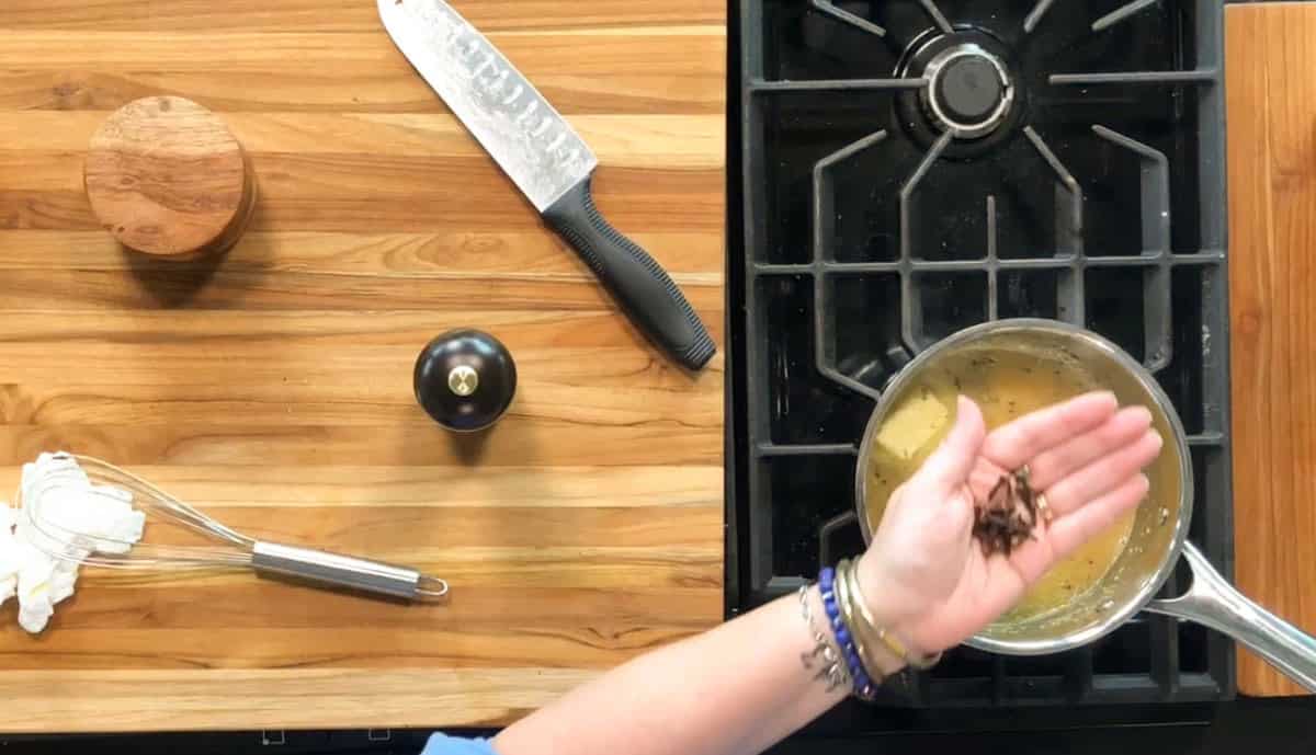 A hand holds several whole cloves above a saucepan with liquid on a stove. Nearby are a chef's knife, a wooden container, a pepper grinder, a whisk, and a towel on a wooden countertop.