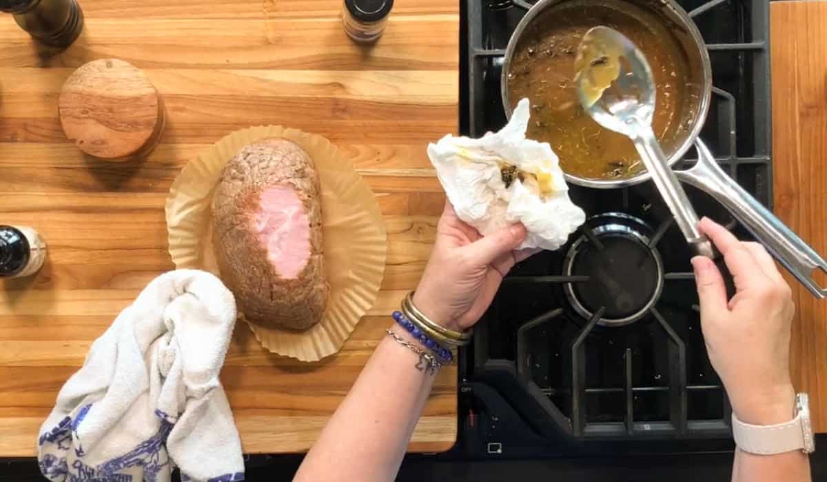 A person wipes a spoon with a napkin over a saucepan of brown liquid on a stove. Nearby on a wooden counter are a cooked beef roast on parchment paper, a towel, and various small containers.
