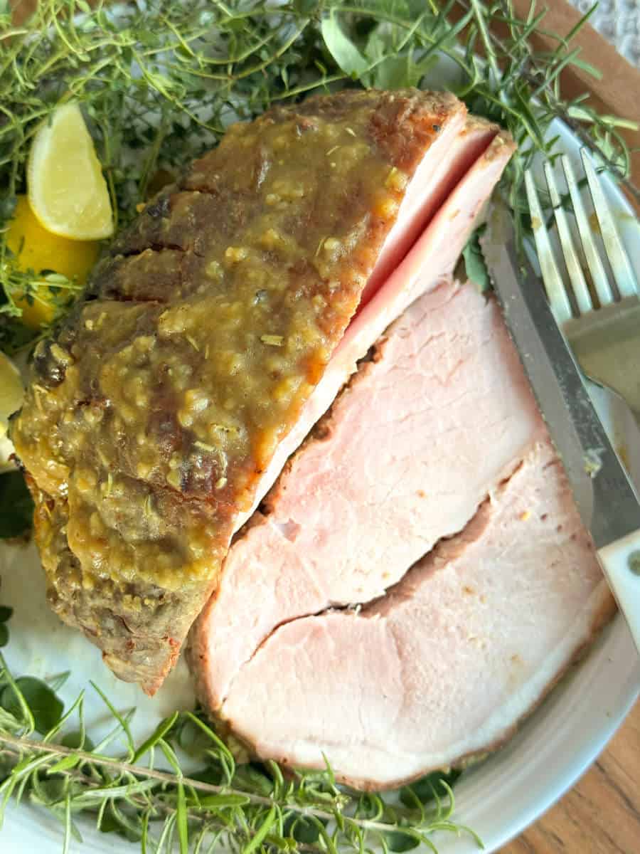 A close-up of two thick slices of baked ham topped with a green sauce on a white plate, garnished with fresh herbs and lemon wedges. A fork and knife are placed to the side of the ham.