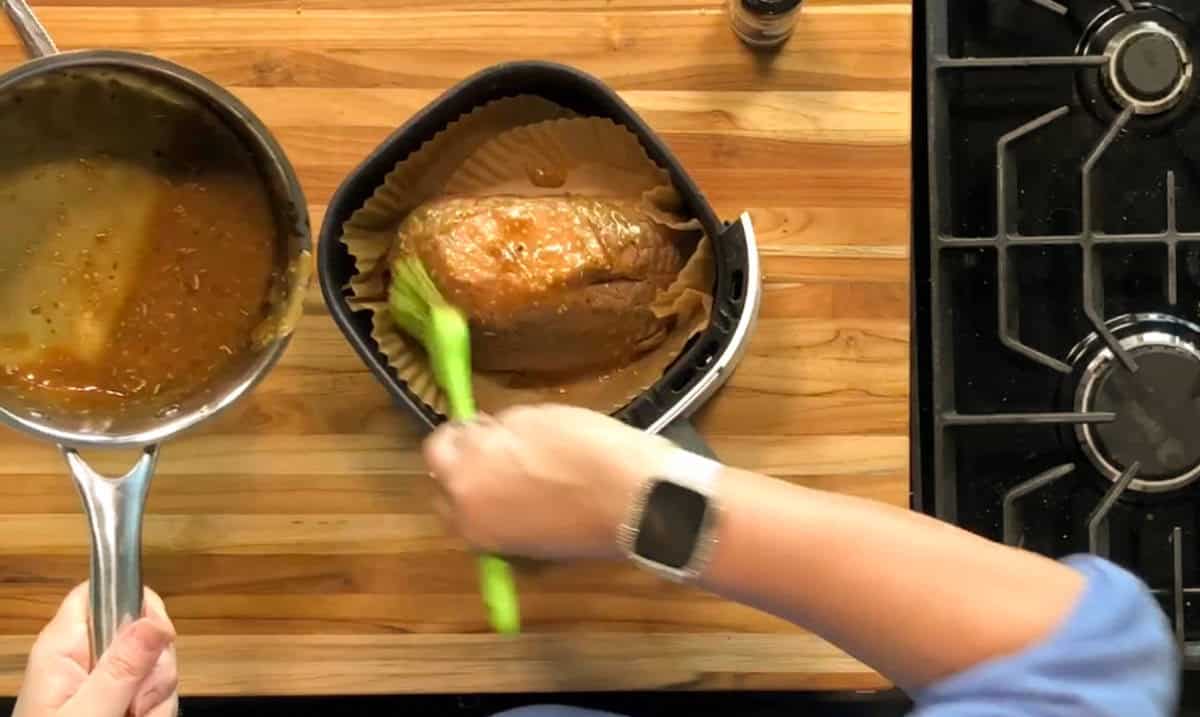 A person brushes sauce onto a roast in an air fryer basket lined with parchment paper. The person holds a pan of sauce in one hand and uses a green brush with the other. A stove and wooden countertop are visible.