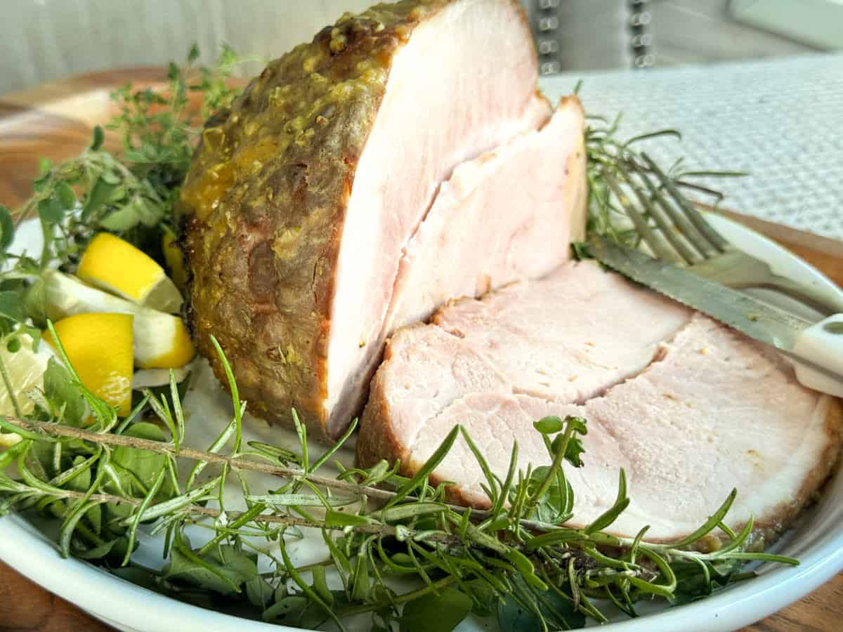 A plate with a thick slice of roasted ham, garnished with fresh herbs and lemon wedges. A knife and fork rest beside the ham, ready for serving.