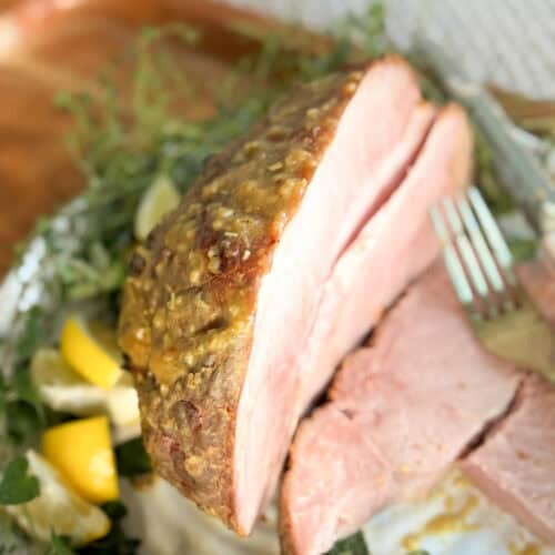 A close-up of a sliced baked ham with a golden crust, garnished with herbs and lemon wedges. A fork rests against the ham, and a couple of slices are lying on the plate.