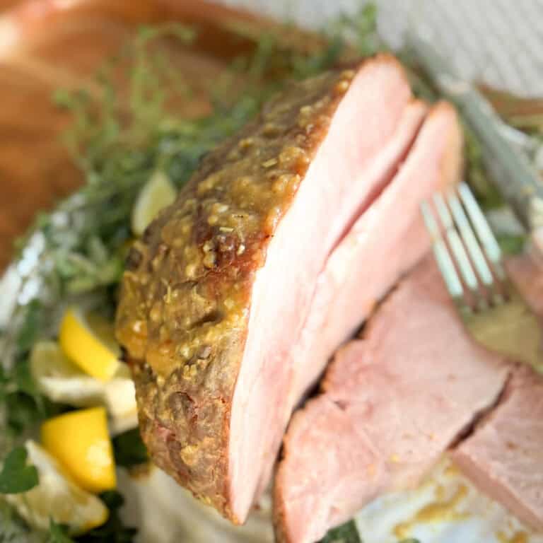 A close-up of a sliced baked ham with a golden crust, garnished with herbs and lemon wedges. A fork rests against the ham, and a couple of slices are lying on the plate.