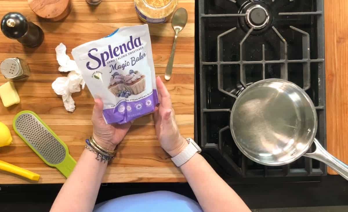 A person holding a bag of Splenda Magic Baker sweetener over a wooden countertop, with a saucepan on a stove, a lemon zester, butter, and spices nearby.