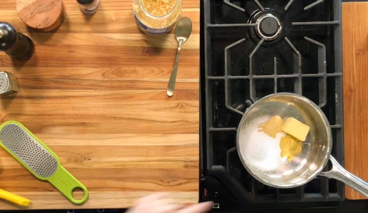 A saucepan on a stove contains butter, mustard, and sugar. A spoon, jar, grater, and various kitchen items are on the wooden counter next to the stove. A hand points toward the saucepan.