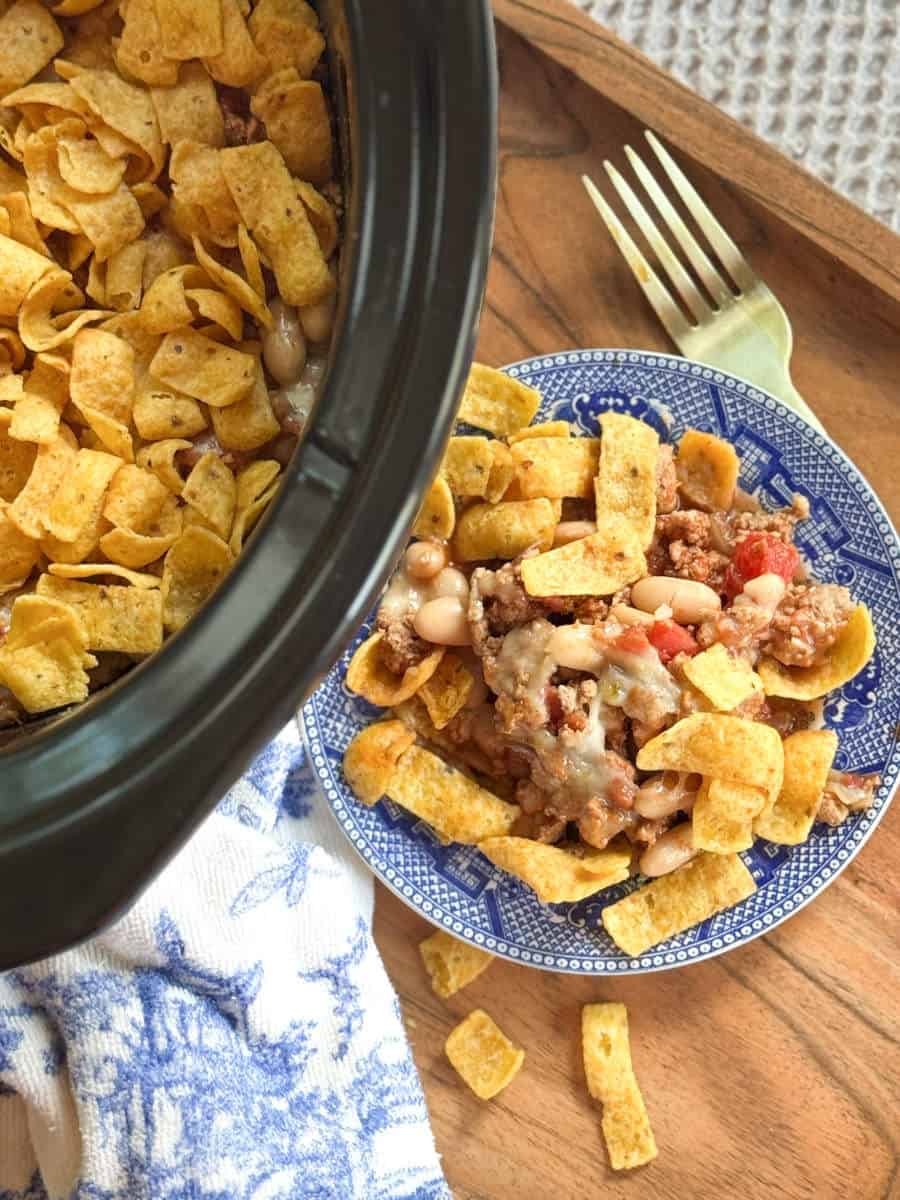 A close-up of a plate with Fritos chips topped with chili, cheese, beans, and tomatoes next to a slow cooker filled with the same dish. A gold fork and scattered chips are on a wooden tray nearby.