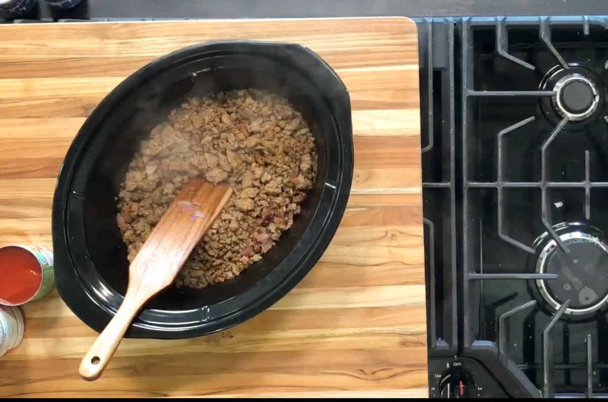 A wooden spatula rests in a black slow cooker filled with cooked ground meat on a wooden countertop next to a stove and a can of tomato sauce.