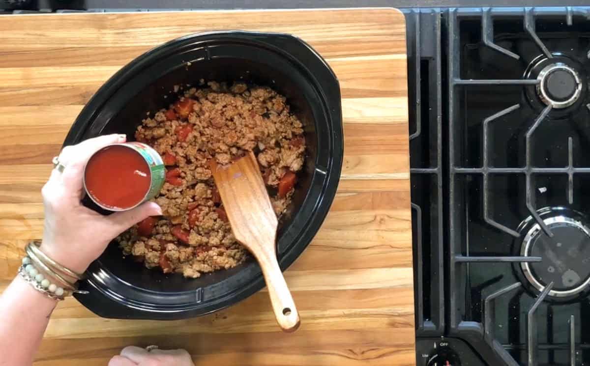 A person pours tomato sauce from a can into a slow cooker containing browned ground meat and diced tomatoes. A wooden spoon rests in the slow cooker, which sits on a wooden countertop next to a stove.