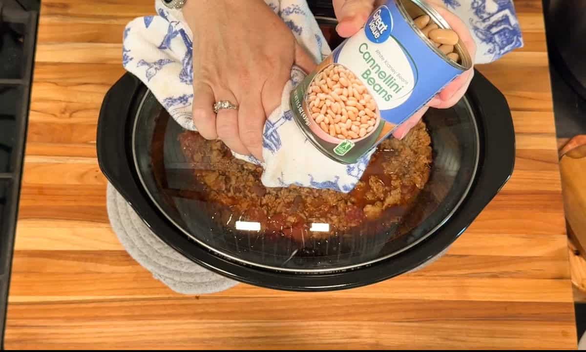 A person holding a towel is opening a can of cannellini beans above a slow cooker filled with cooked ground meat and sauce on a wooden countertop.