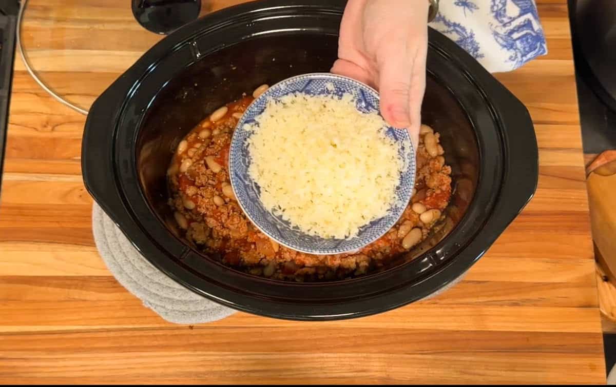 A hand holds a small blue bowl of shredded cheese above a slow cooker filled with chili ingredients, including beans and ground meat, on a wooden countertop.