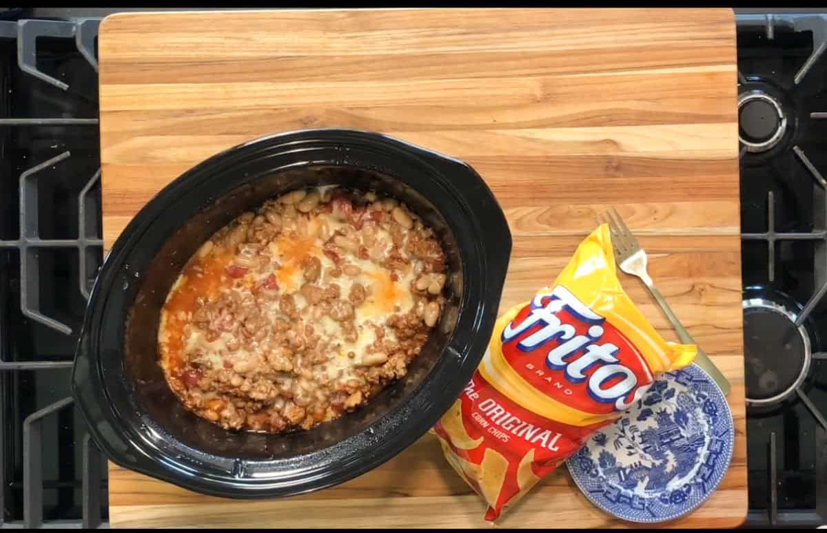 A slow cooker filled with chili sits on a wooden cutting board next to an open bag of Fritos corn chips and a small blue and white patterned bowl. The background shows a stovetop.