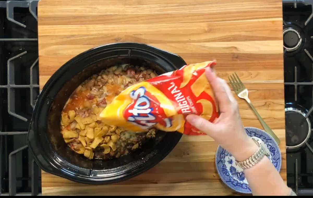 A hand pours Fritos Original corn chips from a bag into a slow cooker filled with a chunky stew or casserole. The slow cooker is on a wooden cutting board next to a blue and white plate with a fork.