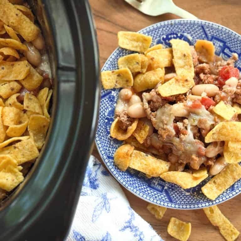 A blue patterned plate with chili, white beans, and corn chips sits beside a slow cooker filled with a similar mixture. Some corn chips are scattered on the wooden table.