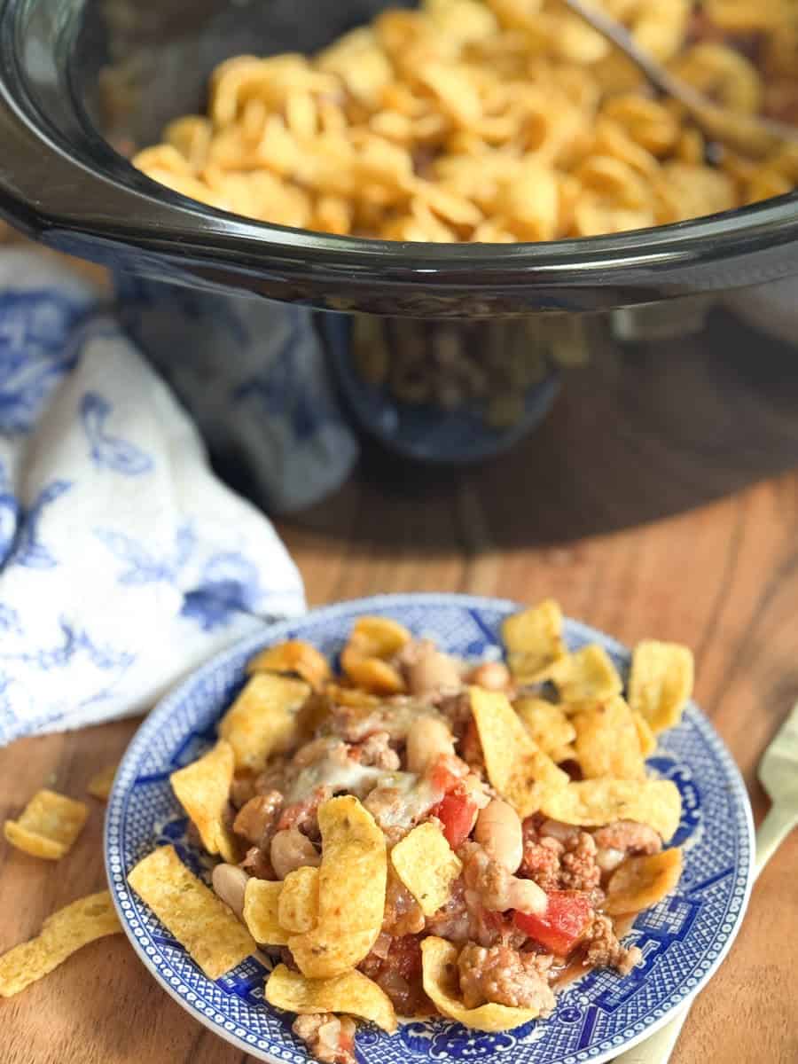 A blue-patterned plate with a serving of chili topped with Fritos corn chips sits on a wooden surface. In the background, a slow cooker filled with more Fritos and a blue-and-white cloth are visible.