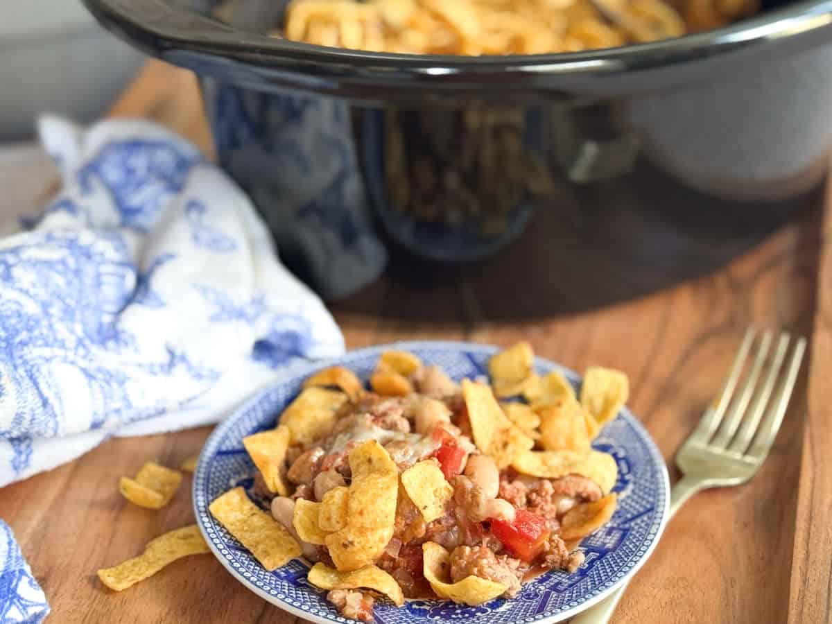 A blue and white plate holds a serving of chili topped with corn chips on a wooden surface, with a fork nearby and a slow cooker with more food in the background. A patterned cloth is partially visible on the left.