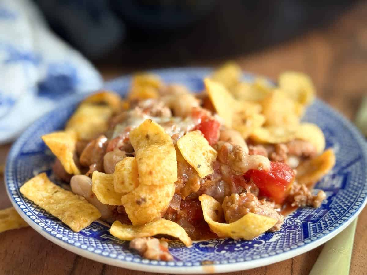A blue and white plate holds a serving of chili topped with corn chips, melted cheese, beans, and diced tomatoes, set on a wooden surface.