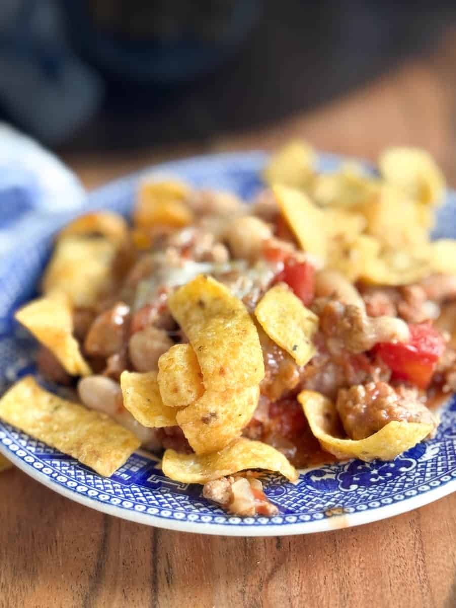 A close-up of a blue patterned plate filled with chili, beans, diced tomatoes, and topped with crispy corn chips, set on a wooden surface.