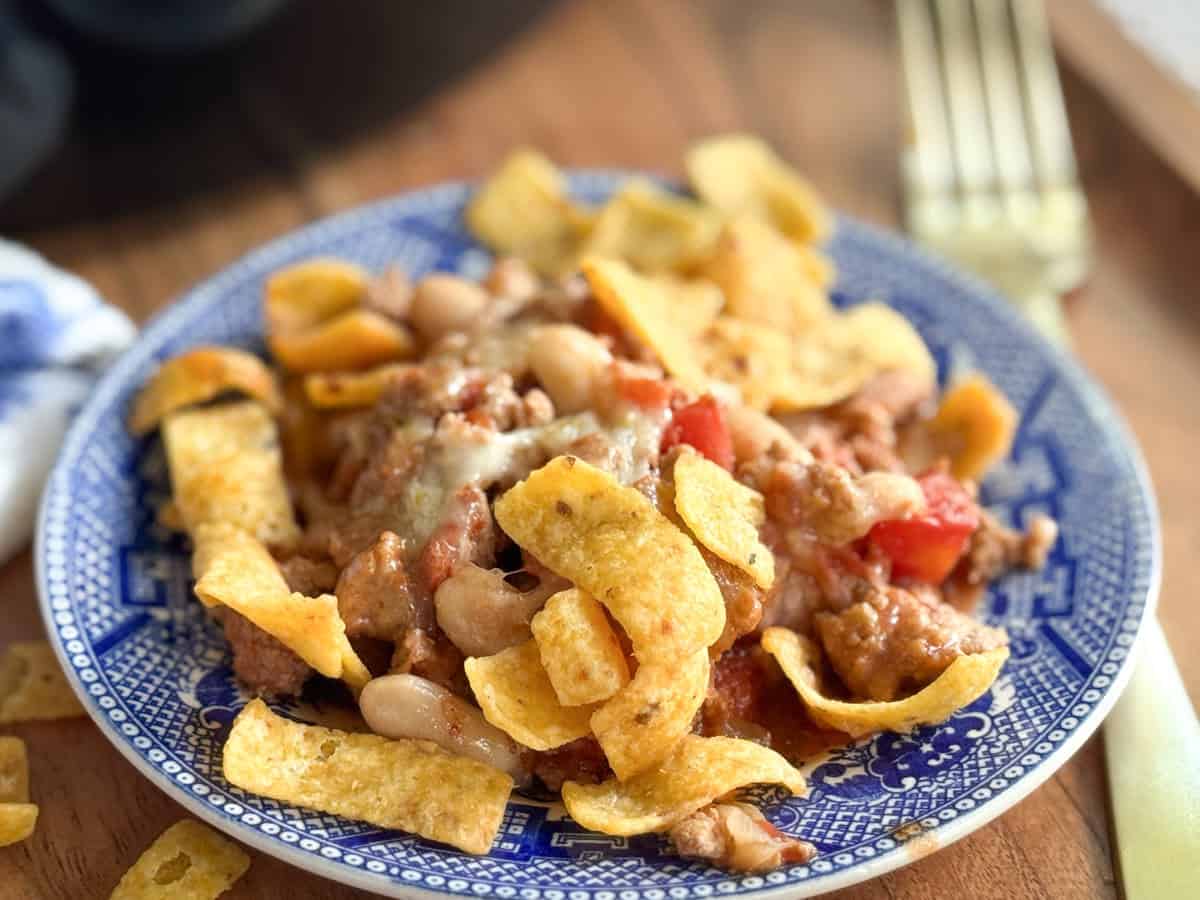 A blue and white plate holds a serving of chili topped with melted cheese, white beans, diced tomatoes, and crunchy corn chips. A fork is visible in the background on a wooden surface.