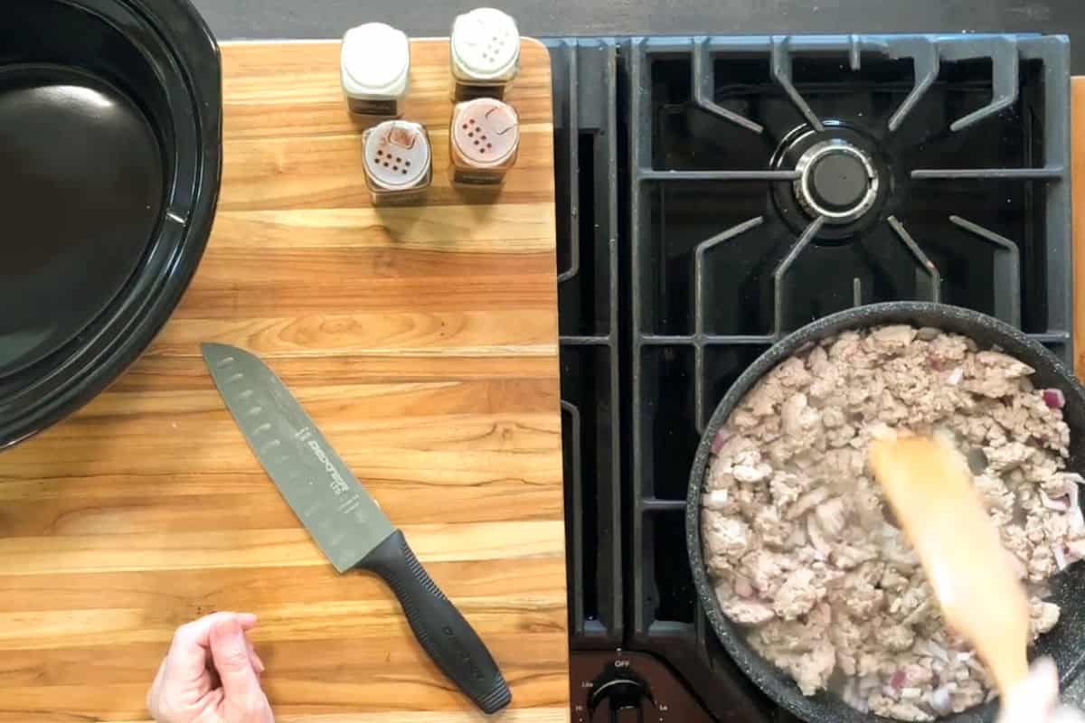 A person holds a wooden spoon, stirring ground meat and onions in a skillet on a gas stove. A chef’s knife, three spice jars, and an empty slow cooker are on a wooden countertop next to the stove.