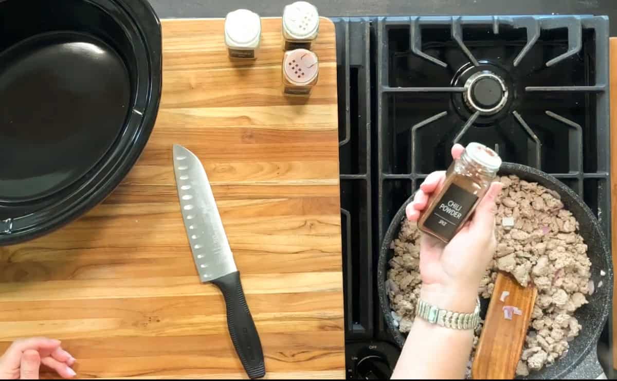 A cutting board with a chef’s knife and spices on the left; on the right, ground meat cooks in a pan on a stove while a hand holds an open container of chili powder above the pan.
