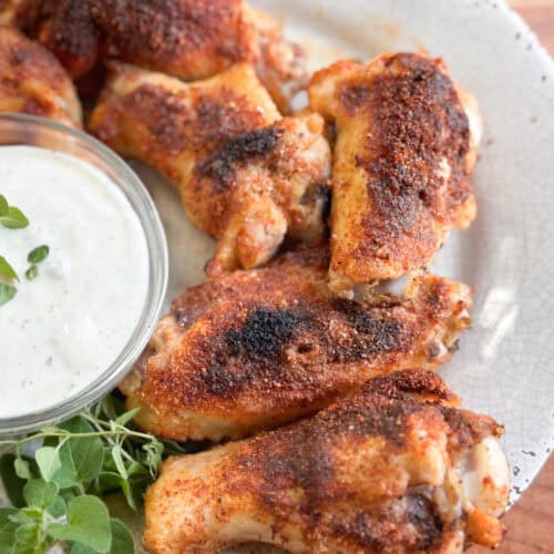 A plate of seasoned and grilled chicken wings is shown next to a small bowl of creamy dipping sauce and a sprig of fresh herbs on a white, textured plate.